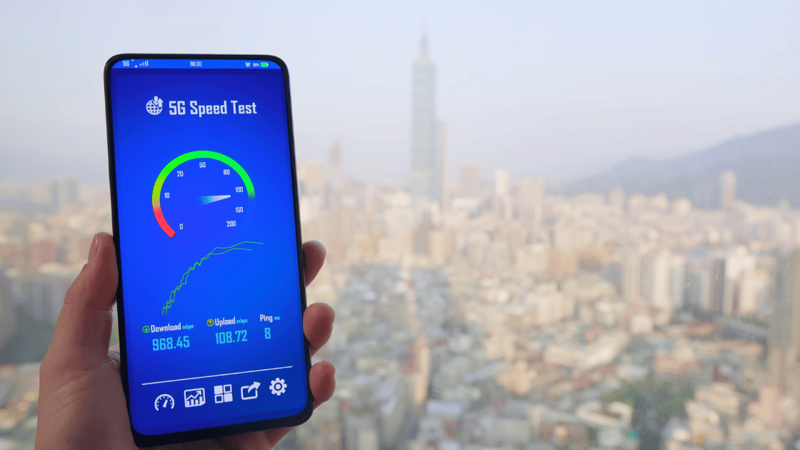 Osoba trzymająca smartfon z otwartą aplikacją do testowania prędkości 5G, pokazującą prędkość pobierania 968,45 Mbps, prędkość wysyłania 108,72 Mbps i ping 8 ms. W tle widoczny jest panoramiczny widok miasta z wysokimi budynkami i mglistym niebem.